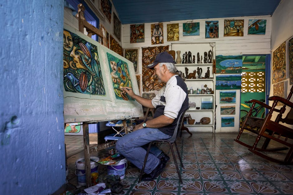 A senior man engaged in painting in a vibrant and colorful art studio filled with paintings and sculptures.